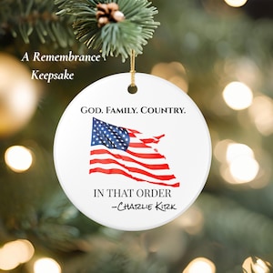 Puede incluir: Adorno redondo de cerámica blanca con una cuerda dorada para colgar. El adorno presenta un diseño de bandera estadounidense ondeando con el texto "GOD. FAMILY. COUNTRY. IN THAT ORDER -Charlie Kirk." Las palabras "A Remembrance Keepsake" también están impresas.