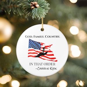 Puede incluir: Adorno navideño redondo y blanco con el texto "GOD. FAMILY. COUNTRY." encima de un diseño de la bandera estadounidense. La bandera es roja, blanca y azul, con una silueta de una mano sosteniendo una cruz. El texto "IN THAT ORDER -Charlie Kirk" está debajo.