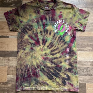 May include: A tie-dye t-shirt with a spiral pattern in shades of green, yellow, and burgundy. The shirt features a graphic on the upper left chest that reads "Grateful Dead" around a lightning bolt and skull design.