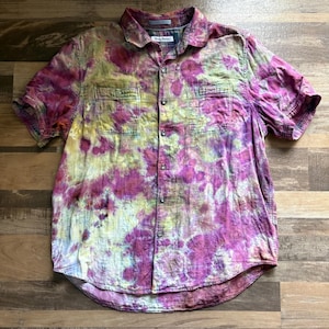 May include: Short-sleeved button-down shirt with a tie-dye pattern in shades of pink, yellow, and green. The shirt has two chest pockets and a collared neckline. The fabric appears to be a lightweight cotton or linen.