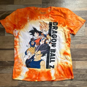 May include: Orange tie-dye t-shirt featuring a graphic of Dragon Ball Z characters. The shirt has the text "DRAGON BALL Z" printed vertically on the right side. The graphic includes multiple characters in various poses and colors.