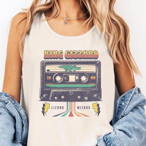 May include: Cream-colored tank top featuring a retro design with the text "KING GIZZARD" above a cassette tape graphic. The cassette has a green lizard illustration and the words "LIZARD" and "WIZARD" below.