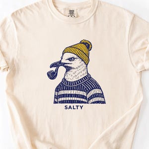 May include: Cream-colored t-shirt featuring a seagull illustration. The seagull is wearing a yellow beanie and a blue and white striped sweater, smoking a pipe. The word "SALTY" is printed below the graphic.