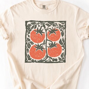 Puede incluir: Camiseta color crema con un dise&ntilde;o cuadrado de cuatro tomates rojos en una vid, enmarcado por hojas y vides verdes. El dise&ntilde;o est&aacute; centrado en la camiseta. La camiseta tiene mangas cortas y cuello redondo.