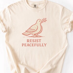 May include: A cream-coloured t-shirt with a dove illustration holding an olive branch, and the text "RESIST PEACEFULLY" beneath. The design is in a muted red-brown hue. The shirt has a crew neck and rolled-up sleeves.