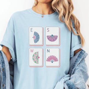 May include: A light blue crew neck t-shirt featuring a graphic print of four rectangular tiles arranged in a square. Each tile displays a letter E, S, W, or N paired with a colorful fan illustration in blue, purple, green, and pink hues.