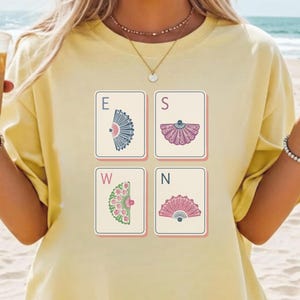 May include: A pale yellow t-shirt featuring a graphic print of four rectangular tiles arranged in a square. Each tile displays a letter E, S, W, or N, paired with a unique illustration of a decorative folding fan in blue, purple, green, or pink.