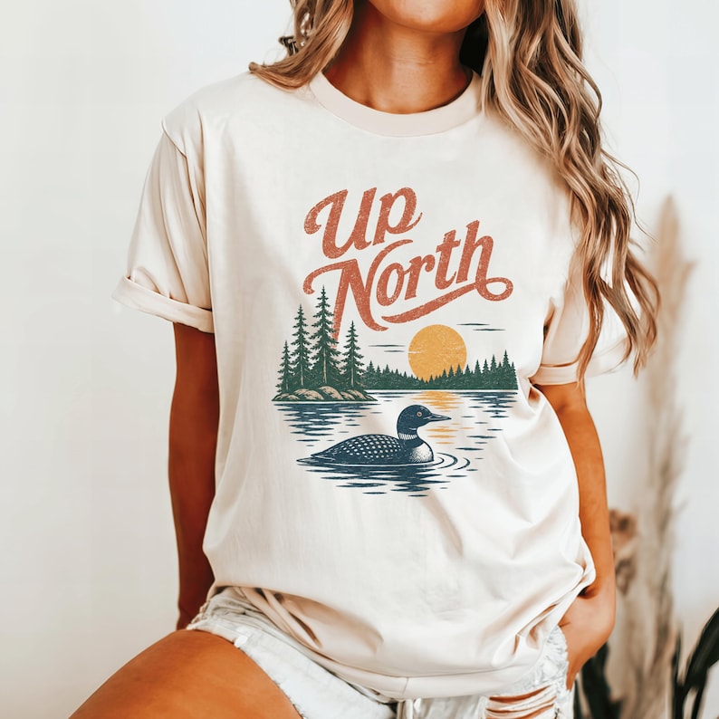 May include: Cream-colored t-shirt featuring a nature-themed graphic. The design includes the text "Up North" in a vintage font, a lake scene with a loon, trees, and a setting sun. The shirt has short sleeves and a crew neck.