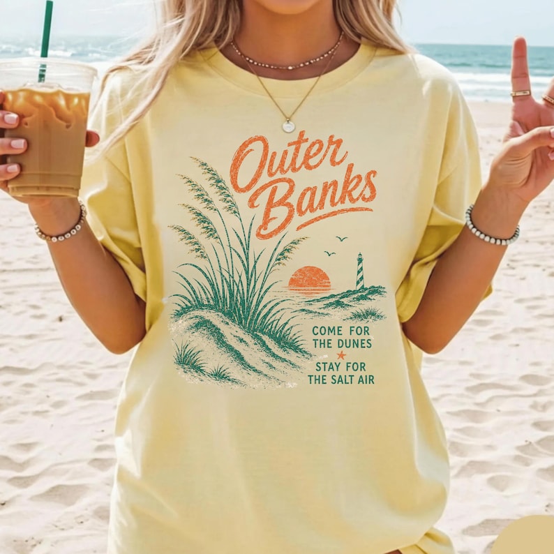 Puede incluir: Una camiseta de manga corta de color amarillo p&aacute;lido con un estampado gr&aacute;fico de dunas de arena, un faro y una puesta de sol. El texto dice Outer Banks, Come for the dunes, Stay for the salt air. El dise&ntilde;o utiliza tinta naranja y verde.