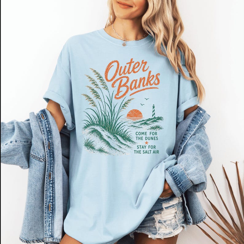 Puede incluir: Una camiseta azul claro con un estampado gr&aacute;fico que incluye el texto Outer Banks, Come for the dunes, Stay for the salt air. El dise&ntilde;o muestra hierba de playa verde, un sol naranja poniente, un faro y dos aves en la costa.