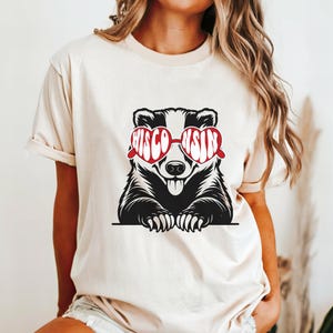 Op de afbeelding: Beige T-shirt met een zwart-witte das-afbeelding met een rode zonnebril met het woord "WISCONSIN" in het wit. De das steekt zijn tong uit. Het shirt heeft korte mouwen.