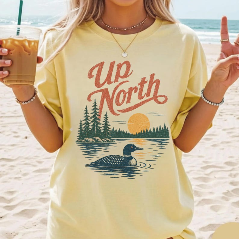 May include: A light yellow t-shirt with the words "Up North" in a retro font. The graphic features a lake scene with a loon, trees, and a setting sun. The shirt is a relaxed fit.