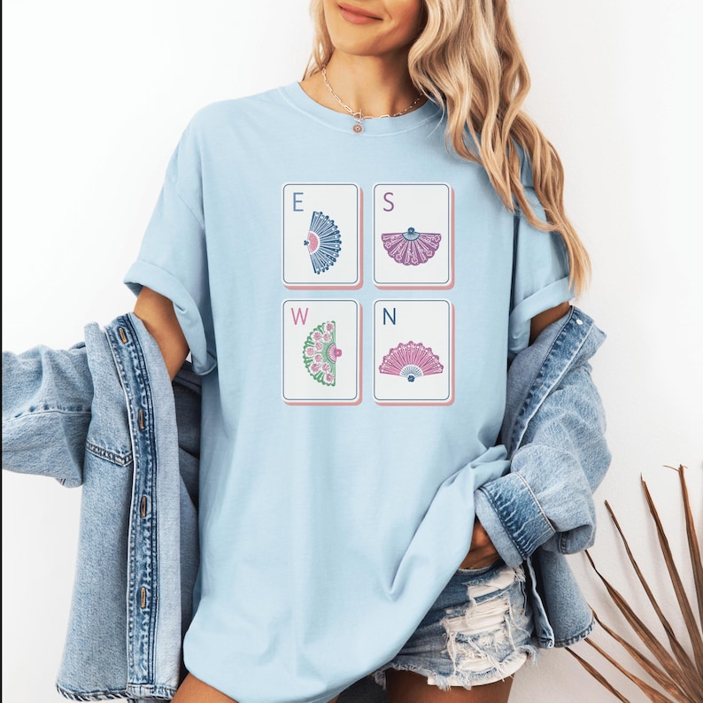 May include: A light blue crew neck t-shirt featuring a graphic print of four rectangular tiles arranged in a square. Each tile displays a letter E, S, W, or N paired with a colorful fan illustration in blue, purple, green, and pink hues.