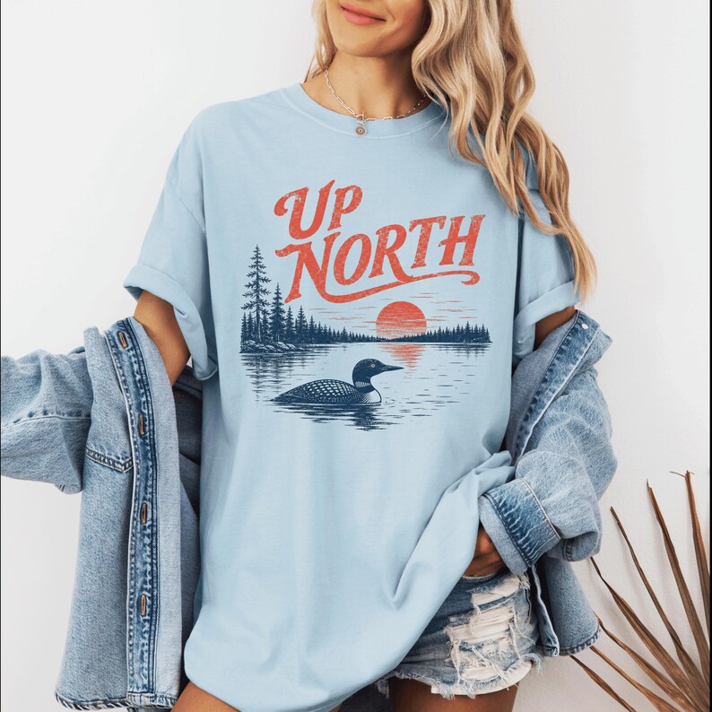 May include: Light blue t-shirt with the words "Up North" in orange script above a graphic of a loon swimming in a lake at sunset. The design includes trees and a sun. The shirt has short sleeves.