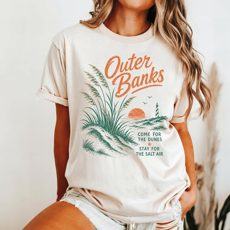 Puede incluir: Una camiseta de manga corta color crema con un gr&aacute;fico de hierba de playa, un faro y una puesta de sol. El texto dice Outer Banks, Come for the dunes, Stay for the salt air. El dise&ntilde;o utiliza tinta de color naranja y verde en el frente.