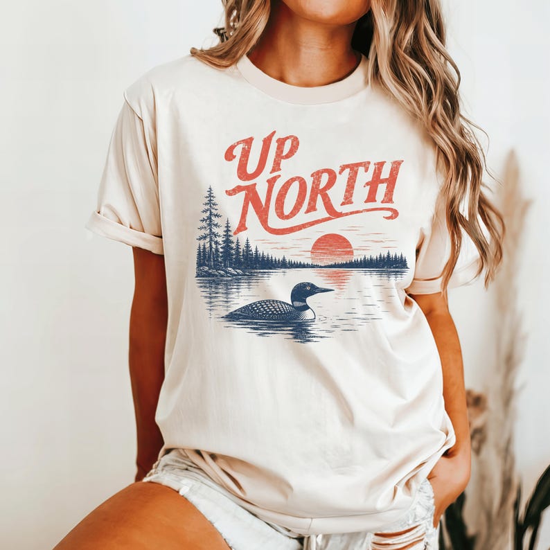 May include: Cream-colored t-shirt featuring a graphic with the text "Up North" in red, above a lake scene with a loon, trees, and a setting sun. The design has a vintage, outdoorsy aesthetic.