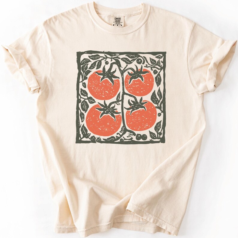 Puede incluir: Camiseta color crema con un dise&ntilde;o cuadrado de cuatro tomates rojos en una vid, enmarcado por hojas y vides verdes. El dise&ntilde;o est&aacute; centrado en la camiseta. La camiseta tiene mangas cortas y cuello redondo.