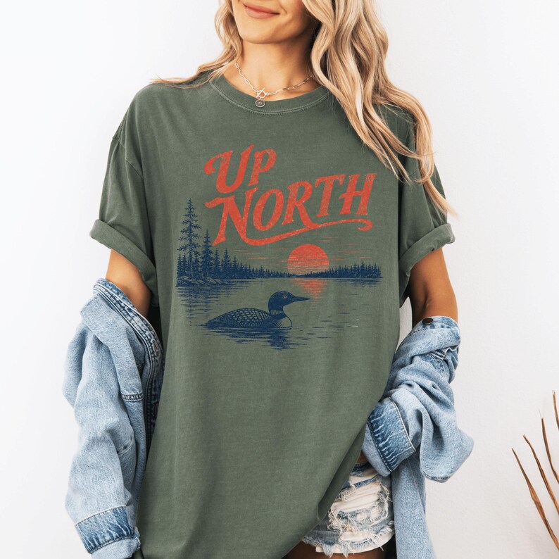 May include: Olive green t-shirt with the words "Up North" in red script. The graphic features a loon on a lake with a sunset and trees. The shirt has rolled-up sleeves and a relaxed fit.