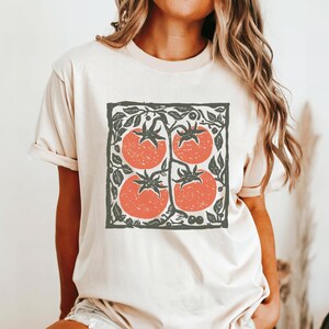Puede incluir: Camiseta color crema con un gr&aacute;fico cuadrado de cuatro tomates rojos con tallos y hojas verdes. El dise&ntilde;o est&aacute; enmarcado por un borde gris oscuro con detalles de hojas. La camiseta tiene mangas cortas.