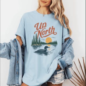 May include: Light blue t-shirt with the text "Up North" in orange script. The graphic features a lake scene with a loon, trees, and a setting sun. The shirt is a relaxed fit.