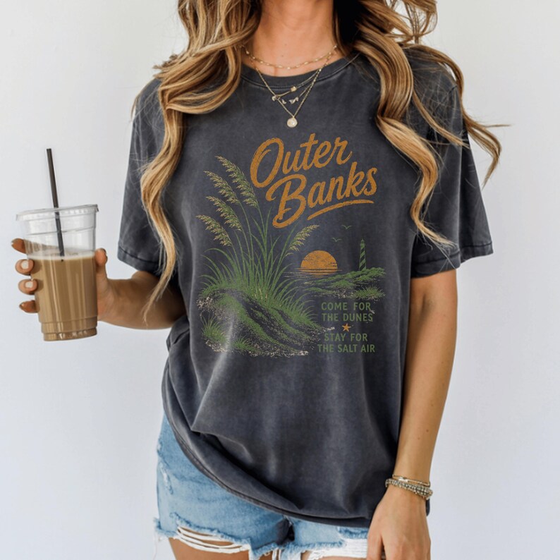 Puede incluir: Una camiseta gris carb&oacute;n de manga corta con un estampado gr&aacute;fico de dunas costeras, un faro y una puesta de sol. El texto dice Outer Banks, Come for the dunes, Stay for the salt air. La camiseta tiene un aspecto desgastado de estilo vintage.