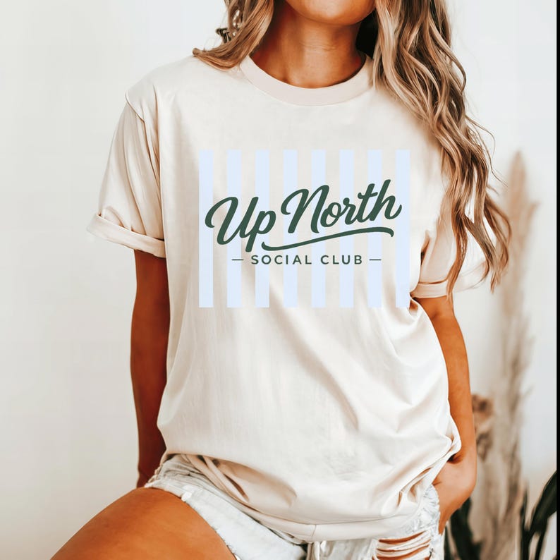 May include: Cream-colored t-shirt with the text "Up North Social Club" in a dark green script font. The text is centered over a light blue striped design. The shirt has short sleeves and a crew neck.