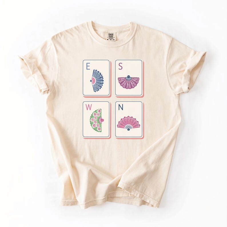 May include: A cream-colored short-sleeve t-shirt featuring a graphic of four rectangular tiles arranged in a square. Each tile displays a letter E, S, W, or N and a unique decorative folding fan illustration in blue, pink, or green color tones.