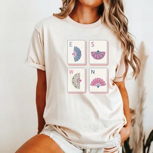 May include: A cream-colored short-sleeve t-shirt featuring a graphic print of four rectangular tiles arranged in a square. Each tile displays a letter E, S, W, or N and a unique decorative fan illustration in blue, purple, green, and pink colors.