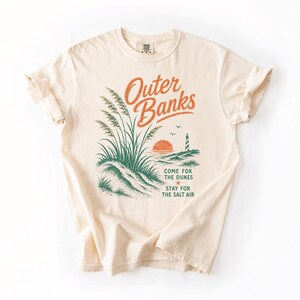 Puede incluir: Una camiseta de manga corta color crema con un estampado gr&aacute;fico de dunas de arena, un faro y una puesta de sol. El texto dice Outer Banks, Come for the dunes, Stay for the salt air. El dise&ntilde;o utiliza tintas de color naranja y verde.
