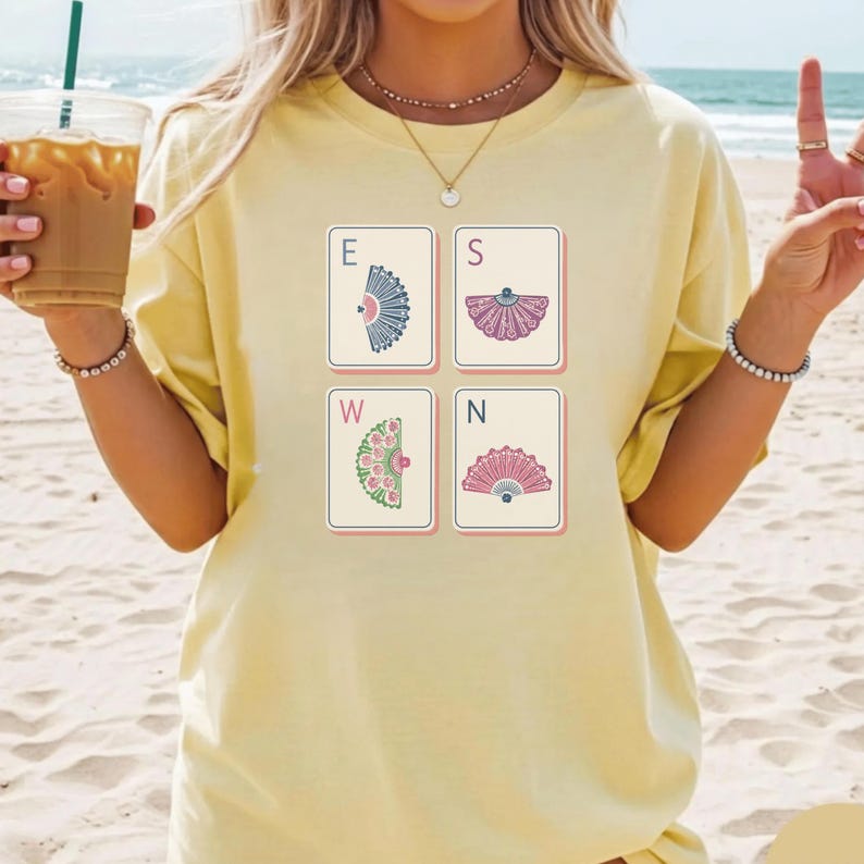 May include: A pale yellow t-shirt featuring a graphic print of four rectangular tiles arranged in a square. Each tile displays a letter E, S, W, or N, paired with a unique illustration of a decorative folding fan in blue, purple, green, or pink.