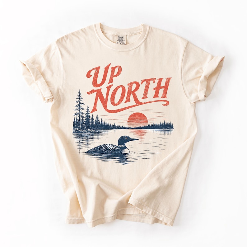 May include: Cream-colored t-shirt featuring a graphic design with the text "UP NORTH" in red, above a lake scene with a loon, trees, and a setting sun. The design is printed on a soft, comfortable tee.