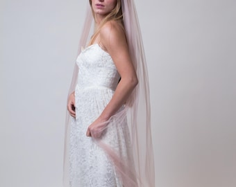 May include: A bride wearing a white lace wedding dress and a long, blush-colored veil. The veil cascades down the back, creating a soft, romantic look. The dress has a fitted bodice and a flowing skirt, perfect for a wedding.