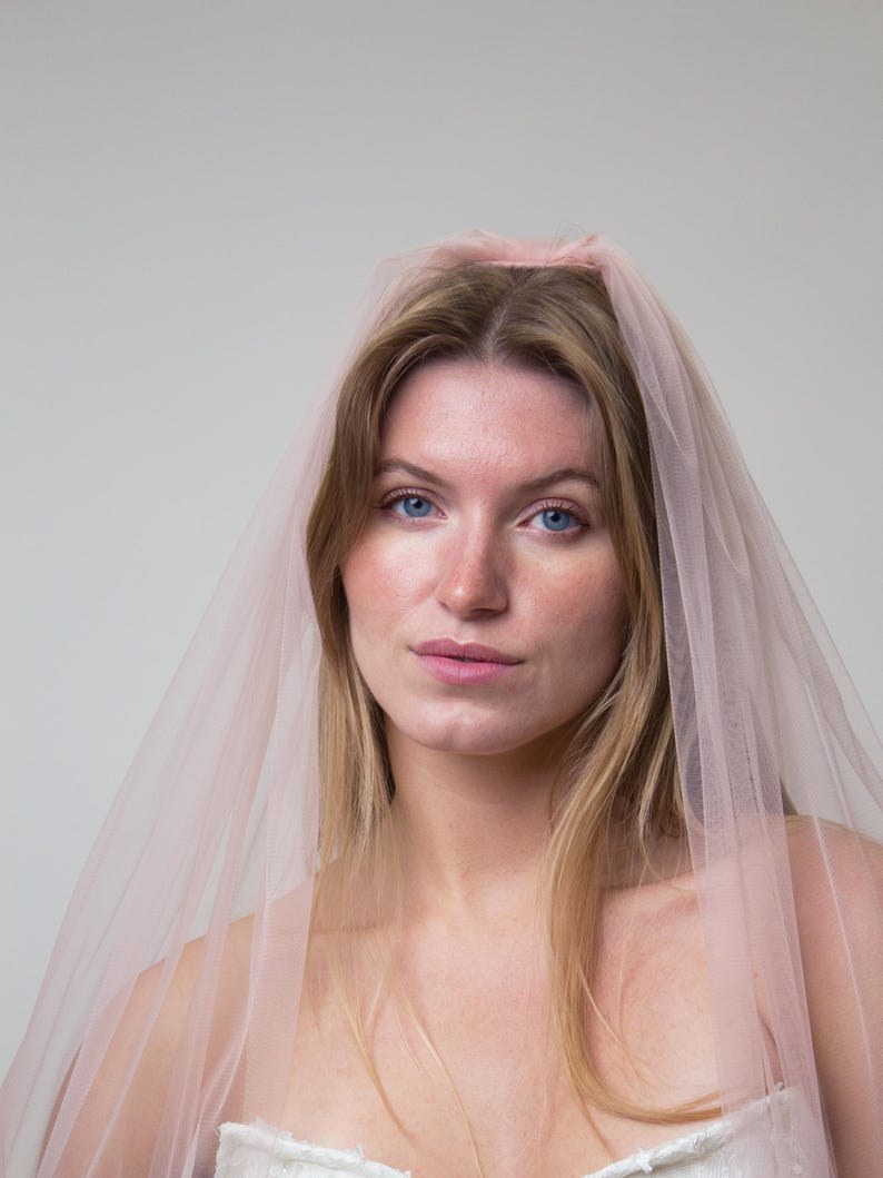 A pale pink tulle bridal veil. The veil is draped over the head and shoulders. The bride has blonde hair and blue eyes. The veil is made of a soft, sheer blush  tulle, creating a delicate and romantic appearance.
