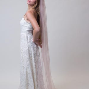 A white lace wedding dress with a long train. A sheer, pale pink veil flows down the back, adding a soft, romantic touch. The dress has a fitted bodice and a full skirt, creating an elegant silhouette.
