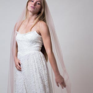 A white lace wedding dress with a sweetheart neckline and a flowing, floor-length skirt. A sheer, blush-coloured veil drapes over the shoulders. The dress and veil create a romantic and elegant bridal look.