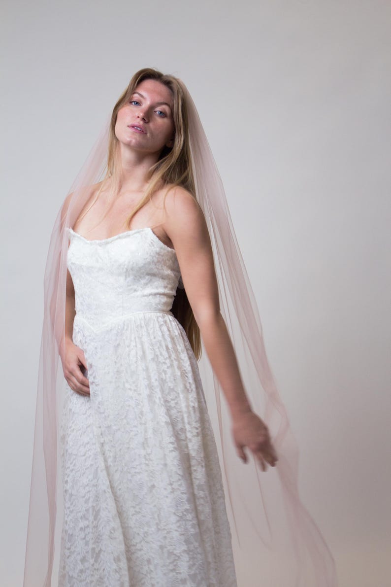 A white lace wedding dress with a sweetheart neckline and a flowing, floor-length skirt. A sheer, blush-coloured veil drapes over the shoulders. The dress and veil create a romantic and elegant bridal look.