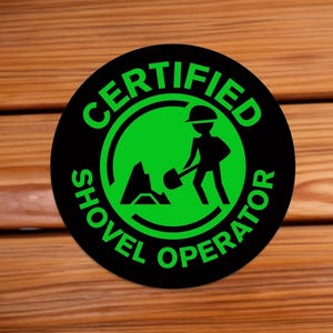 May include: A black and green round sticker with the text "CERTIFIED SHOVEL OPERATOR" in a white circle. A green silhouette of a person with a shovel is inside the circle.