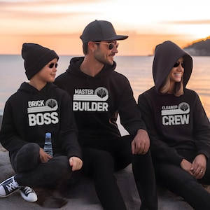 Puede incluir: Una familia lleva sudaderas con capucha negras con texto blanco y un gráfico de un ladrillo Lego. El texto en las sudaderas con capucha dice "Brick Boss", "Master Builder" y "Cleanup Crew".