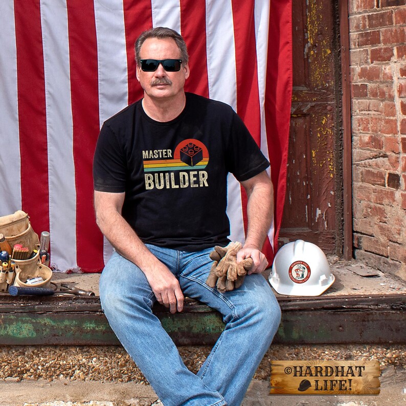 May include: A black short-sleeved t-shirt featuring a graphic print with the text MASTER BUILDER and a building block icon in orange, yellow, and green stripes. The shirt is worn with blue denim jeans, dark sunglasses, and tan work gloves.