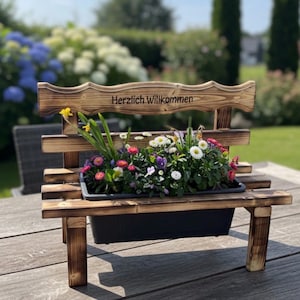 Puede incluir: Una jardinera en forma de banco de madera con un contenedor de pl&aacute;stico negro lleno de flores coloridas. El banco tiene un respaldo con la inscripci&oacute;n "Herzlich Willkommen" y est&aacute; sobre una superficie de madera.