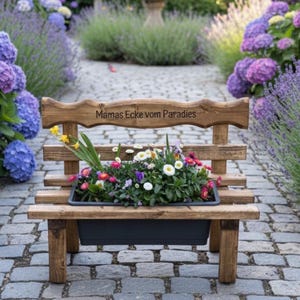 Puede incluir: Un banco de madera r&uacute;stico con una jardinera llena de flores coloridas. El banco tiene la inscripci&oacute;n "Mamas Ecke vom Paradies". Est&aacute; situado en un camino empedrado, rodeado de hortensias y otras plantas, creando una encantadora escena de jard&iacute;n.