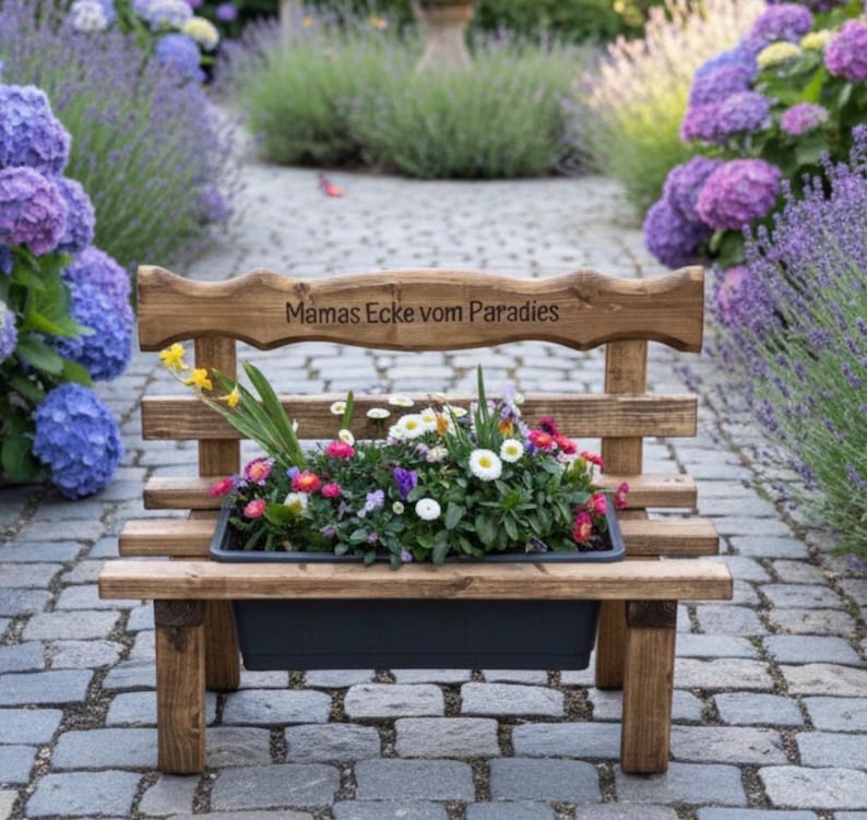 Puede incluir: Un banco de madera r&uacute;stico con una jardinera llena de flores coloridas. El banco tiene la inscripci&oacute;n "Mamas Ecke vom Paradies". Est&aacute; situado en un camino empedrado, rodeado de hortensias y otras plantas, creando una encantadora escena de jard&iacute;n.