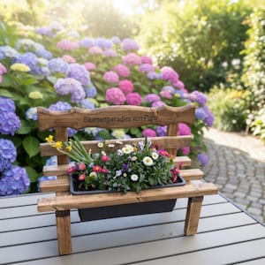Puede incluir: Un peque&ntilde;o banco de madera con una jardinera llena de flores coloridas. El banco tiene un dise&ntilde;o r&uacute;stico y el texto "Blumenparadies von Papa" est&aacute; tallado en el respaldo. El banco est&aacute; sobre una mesa frente a un fondo de hortensias florecientes.