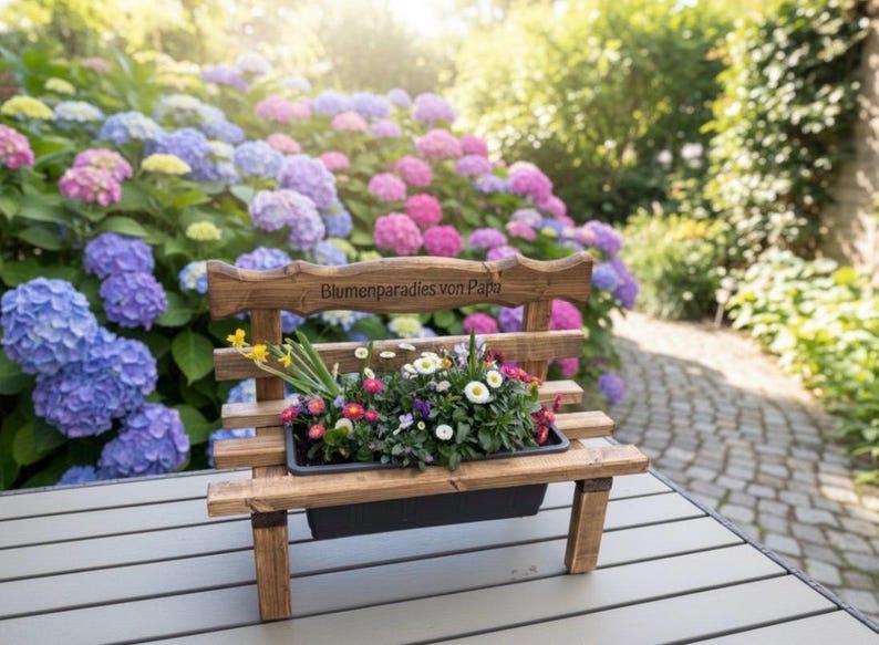 Puede incluir: Un peque&ntilde;o banco de madera con una jardinera llena de flores coloridas. El banco tiene un dise&ntilde;o r&uacute;stico y el texto "Blumenparadies von Papa" est&aacute; tallado en el respaldo. El banco est&aacute; sobre una mesa frente a un fondo de hortensias florecientes.