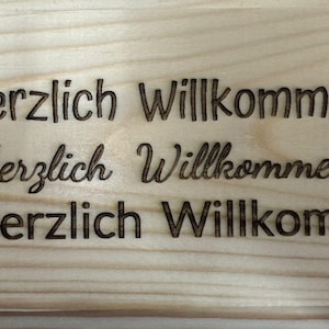 Puede incluir: Un letrero de madera con las palabras "1. Herzlich Willkommen", "2. Herzlich Willkommen" y "3. Herzlich Willkommen" grabadas. El letrero est&aacute; hecho de madera clara con vetas visibles.