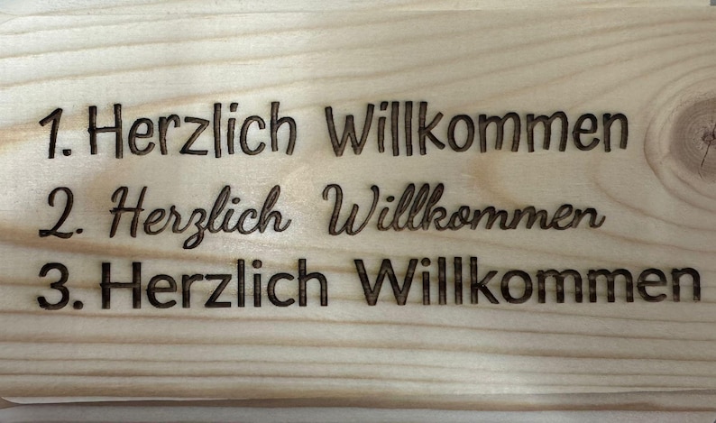 Puede incluir: Un letrero de madera con las palabras "1. Herzlich Willkommen", "2. Herzlich Willkommen" y "3. Herzlich Willkommen" grabadas. El letrero est&aacute; hecho de madera clara con vetas visibles.