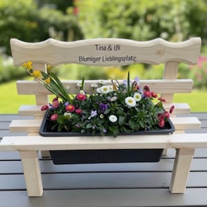 Puede incluir: Un peque&ntilde;o banco de madera para plantas, lleno de flores coloridas. El banco tiene un respaldo decorativo con el texto "Tina & Uli" y "Blumiger Lieblingsplatz". La jardinera est&aacute; sobre una mesa gris, con un fondo verde borroso.