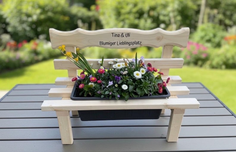 Puede incluir: Un peque&ntilde;o banco de madera para plantas, lleno de flores coloridas. El banco tiene un respaldo decorativo con el texto "Tina & Uli" y "Blumiger Lieblingsplatz". La jardinera est&aacute; sobre una mesa gris, con un fondo verde borroso.
