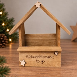 Könnte beinhalten: Eine hellbraune Holzkonstruktion in Form eines kleinen Hauses mit offener Vorderseite und Dach. Die Frontplatte trägt den eingravierten Text "Weihnachtsmarkt to go" und ist mit drei kleinen, am Rahmen befestigten Holzsternen verziert.