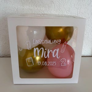 Könnte beinhalten: Eine weiße Box mit einem transparenten Fenster, das vier Ballons enthält. Die Ballons sind weiß, gold und rosa. Auf der Box steht "Einschulung Mira 19.08.2025" mit Illustrationen zum Thema Schule.
